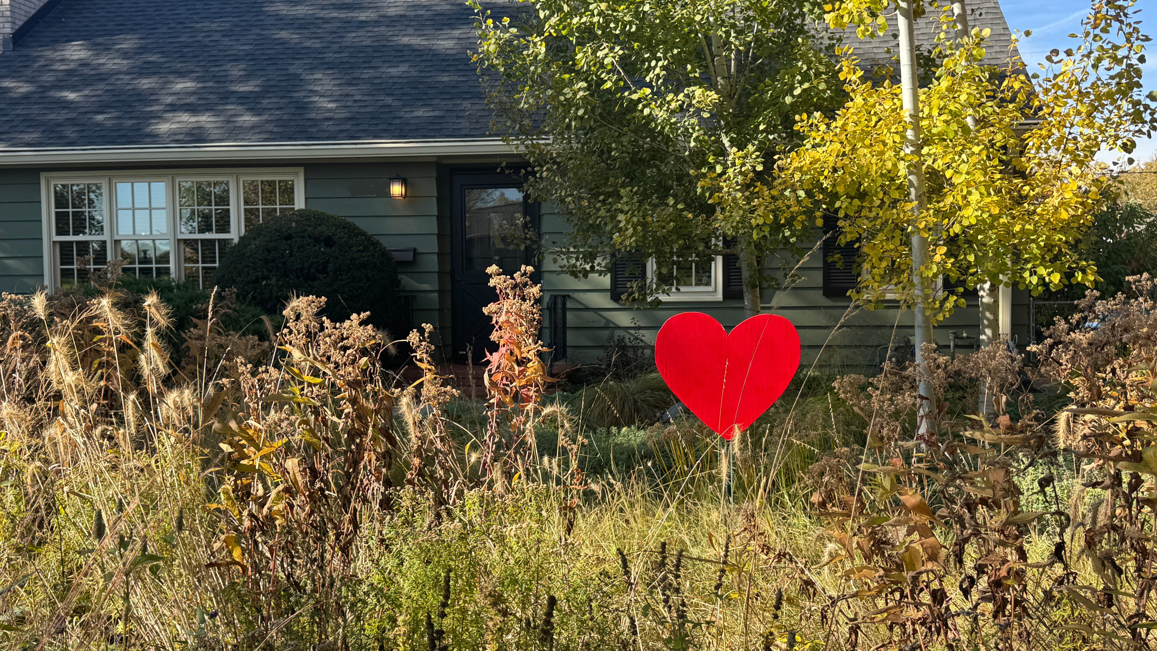 Red heart-shaped sign in front of a house with trees and bushes.